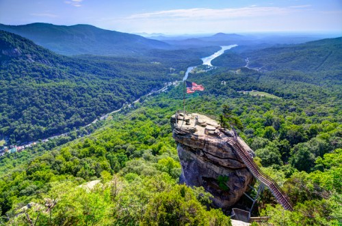 A scenic view from a rocky overlook, featuring lush green mountains and a river, with an American flag on a rock ledge.