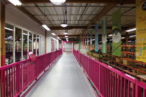 A brightly colored walkway with pink railings, leading through a spacious warehouse filled with shelves and banners.