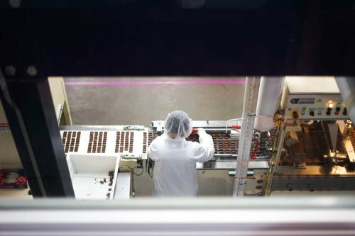 A worker in a hairnet and lab coat operates machinery in a chocolate production facility.