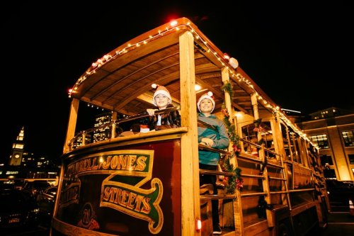 Two people in Santa hats smile from a festive, decorated trolley at night, surrounded by city lights.