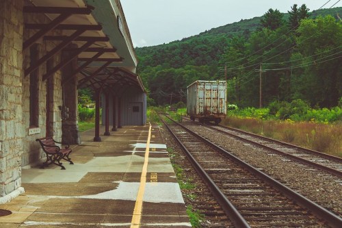Abandoned train station platform with a bench, overlooking railway tracks and a freight car against a green mountain backdrop.