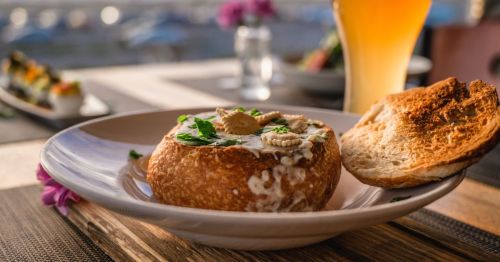 photo of clam chowder in a bread bowl on a white plate next to a glass of beer on a restaurant patio