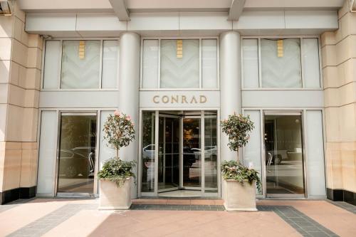 Entrance of the Conrad hotel, featuring large glass doors and decorative potted plants on either side.
