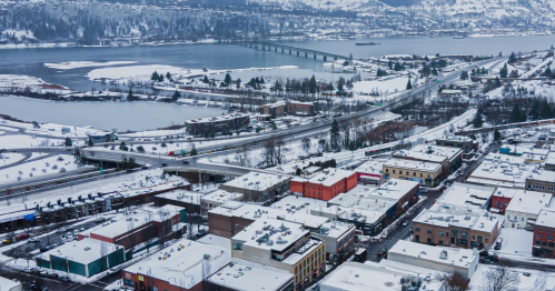 Aerial view of a snowy town with a river, bridge, and mountains in the background, showcasing winter scenery.