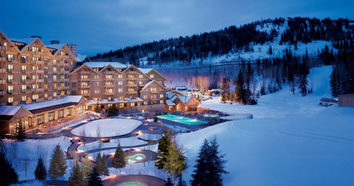 A snowy resort at dusk, featuring illuminated buildings and outdoor pools surrounded by trees and mountains.