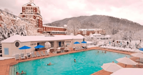 A snowy landscape surrounds a heated outdoor pool with people swimming, set against a historic hotel and mountains.
