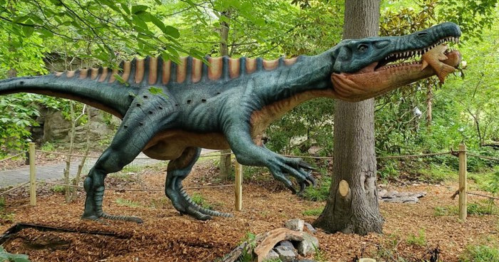 A large dinosaur sculpture stands in a wooded area, holding a smaller dinosaur in its mouth.