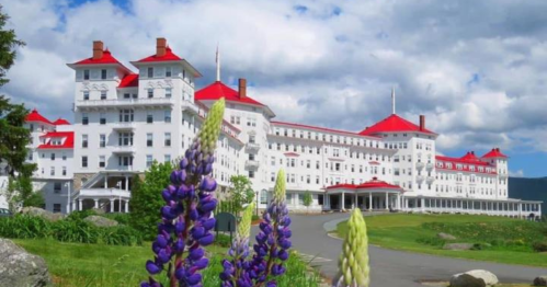A large white hotel with red roofs surrounded by green grass and colorful flowers under a blue sky with clouds.