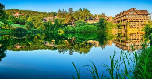 Scenic view of a calm lake reflecting lush greenery and rustic buildings under a clear blue sky.
