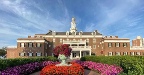 Historic brick building with a tower, surrounded by vibrant flower beds and a blue sky.