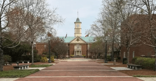 A brick pathway leads to a clock tower building, surrounded by trees and benches in a serene campus setting.
