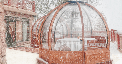 A cozy outdoor dining pod surrounded by snow, with a table set inside and a scenic winter backdrop.