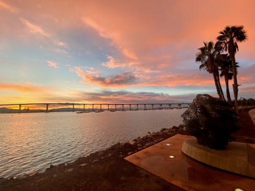 A vibrant sunset over a calm bay, with palm trees and a bridge in the background, and a sculpture in the foreground.