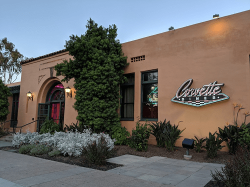 Exterior of the Corvette Diner, featuring a colorful sign and lush greenery in front of a peach-colored building.
