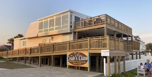 A wooden restaurant with a large deck, "Crab's Claw," located near the beach, featuring a sign and outdoor seating.