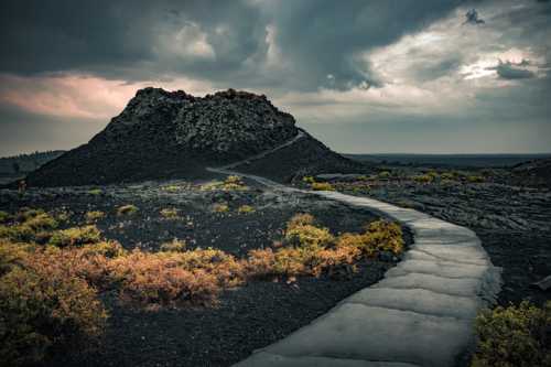 A winding path leads through a volcanic landscape with dark rocks and sparse vegetation under a cloudy sky.