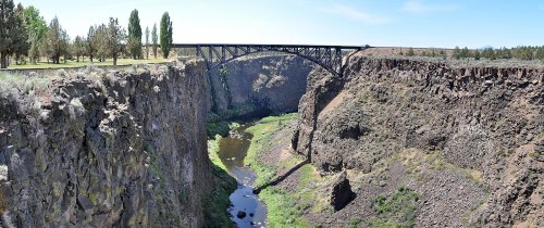 A panoramic view of a deep canyon with a river below and a bridge spanning across the top. Lush greenery is visible.