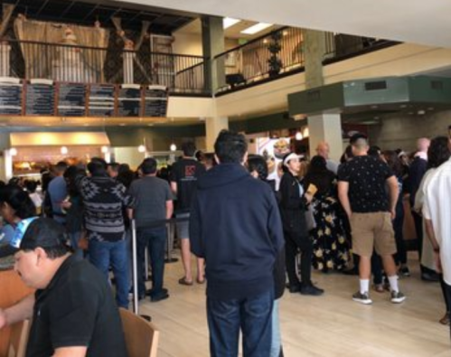 A crowded restaurant interior with people waiting in line and dining, featuring a two-level seating area.