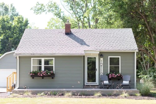 A small gray house with a front porch, flower boxes, and two chairs, surrounded by trees and greenery.