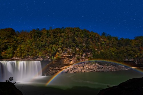 A serene waterfall under a starry sky, with a vibrant rainbow arching over the calm water below.