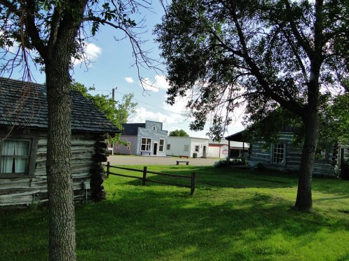 A grassy area with two log cabins and a modern building in the background, framed by trees under a blue sky.