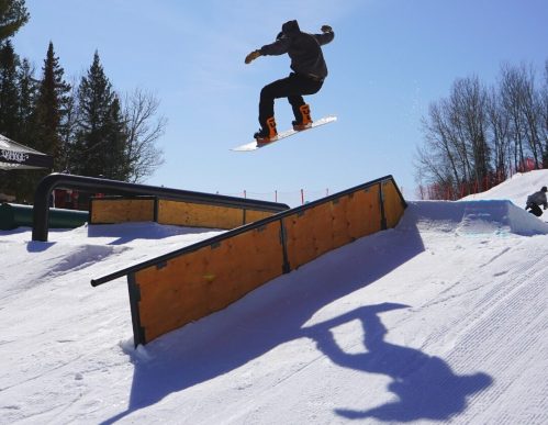 A snowboarder jumps over a rail in a snowy terrain park under a clear blue sky. Trees are visible in the background.
