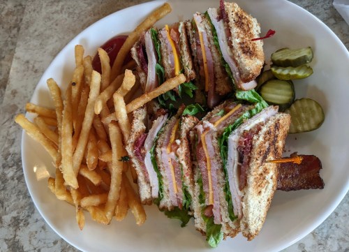 A plate with a stacked club sandwich, crispy fries, and pickles on the side.