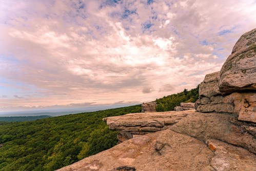 A rocky outcrop overlooks a lush green forest under a cloudy sky, with distant hills visible on the horizon.