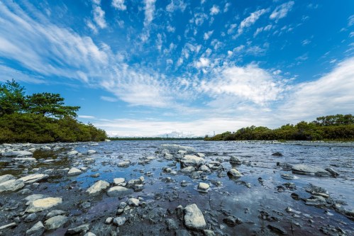 A rocky riverbed under a blue sky with scattered clouds, surrounded by greenery and distant industrial structures.