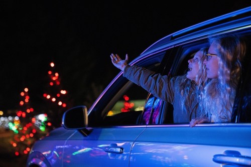 Two women in a car, enjoying colorful holiday lights outside, with one gesturing excitedly.