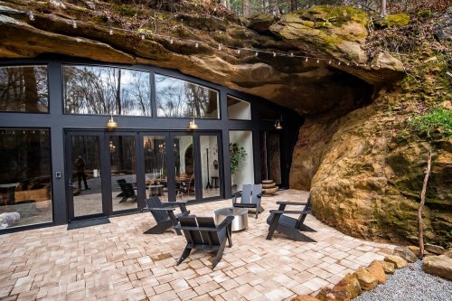 A modern outdoor patio with black chairs, set against a large rock formation and surrounded by trees.
