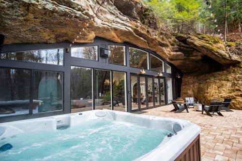 A hot tub in front of a modern glass-walled cabin set against a rocky cliff in a wooded area.