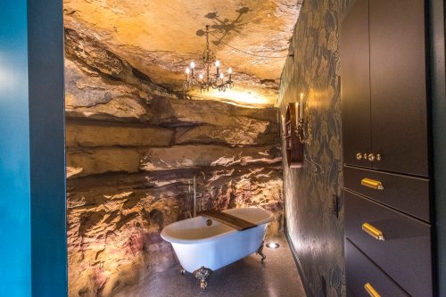 A unique bathroom featuring a freestanding tub against a rock wall, illuminated by a chandelier and soft lighting.
