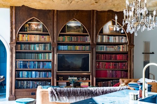 A wooden bookshelf with arched shelves filled with colorful books, framed art above, and a cozy living space below.
