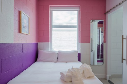 A cozy bedroom with pink and purple walls, a large window, and a neatly made bed with white towels.