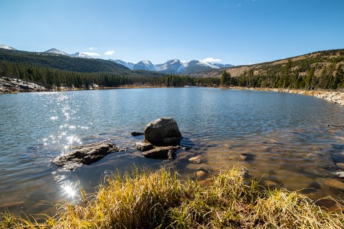 A serene lake surrounded by mountains, with sparkling water and grassy shores under a clear blue sky.