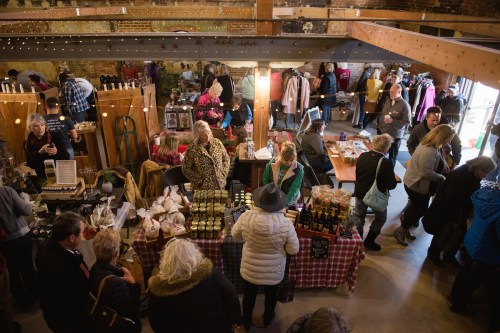 A bustling market scene with vendors, shoppers, and various goods displayed on tables in a cozy indoor space.
