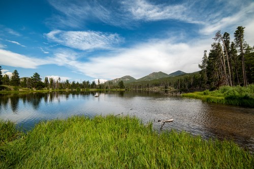 A serene lake surrounded by lush greenery and mountains under a blue sky with scattered clouds.