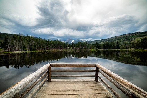 A wooden dock extends over a calm lake, surrounded by trees and mountains under a cloudy sky.
