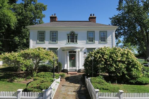 A charming white two-story house with green shutters, surrounded by lush greenery and a white picket fence.