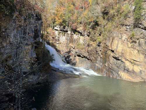 A serene waterfall cascades into a calm pool, surrounded by autumn foliage and rocky cliffs.