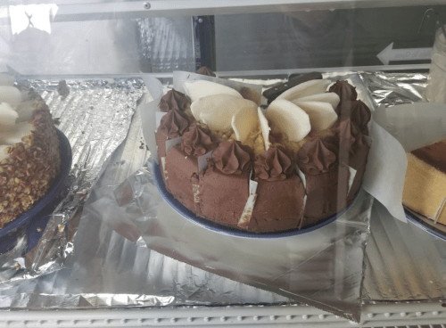 A chocolate cake topped with banana slices and chocolate frosting, displayed in a glass case with other desserts.