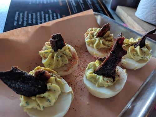 Five deviled eggs topped with crispy bacon and sprinkled with paprika on a tray.