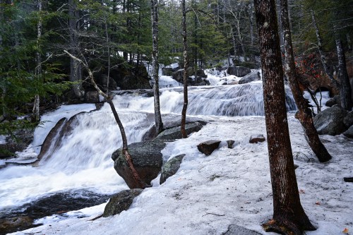 A serene waterfall cascades over rocks, surrounded by trees and patches of snow in a tranquil forest setting.