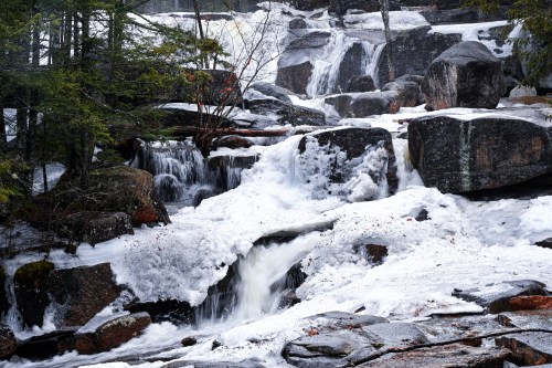 A serene waterfall cascades over rocky terrain, surrounded by lush greenery and frothy white water.