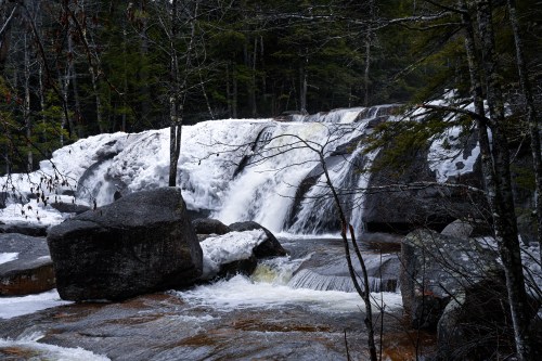 A serene waterfall cascades over rocks, surrounded by snow and lush green trees in a tranquil forest setting.