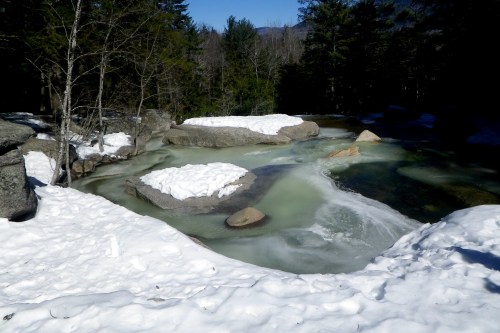 A serene winter scene featuring a partially frozen river surrounded by snow-covered rocks and trees.
