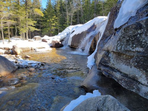 A serene stream flows through a snowy landscape, surrounded by rocks and trees in a peaceful forest setting.