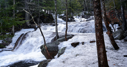 A serene scene of a flowing river over rocks, surrounded by trees and patches of snow.