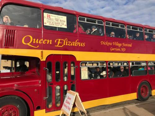 A red and yellow double-decker bus named "Queen Elizabus" with passengers, parked at the Dickens Village Festival.